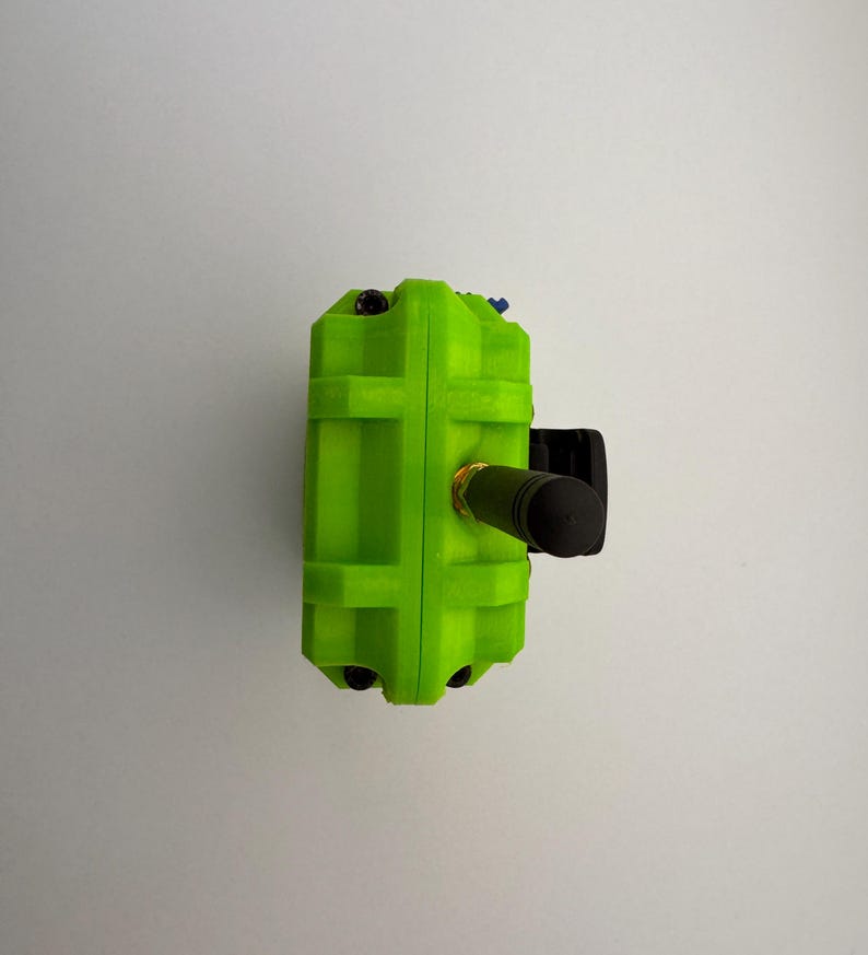 May include: A bright green rectangular device with a black cylindrical component extending from the side. The device has a textured surface and small black and blue accents. The background is a plain white wall.