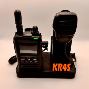 May include: Black Yaesu FT5D handheld radio with a speaker microphone in a black charging base. The radio has a small screen and several buttons. The base has the text "KR4S" in orange.