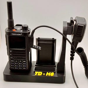 May include: Black Tidradio TD-H8 two-way radio with a charging base and microphone. The radio has a screen, buttons, and an antenna. The charging base has a slot for a battery. The microphone is connected with a coiled cable. The text "TD-H8" is visible.