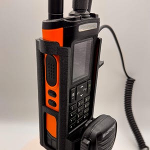 May include: Black and orange Baofeng two-way radio with a black microphone. The radio has a screen, a keypad, and an antenna. The radio is in a black holder. The microphone is connected to the radio with a black cord.