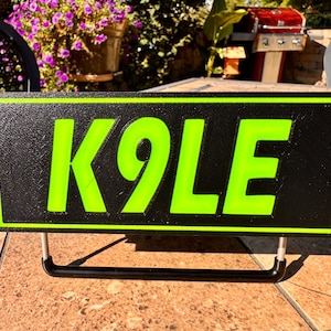 May include: Black rectangular sign with the word "K9OLE" in bright green lettering. The sign has a black border and is mounted on a black metal stand. The sign is outdoors on a tiled surface.