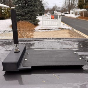 May include: A black antenna with a rectangular base and a textured, dark gray panel. The antenna is mounted on a vehicle's surface, with a residential street and snowy landscape in the background.