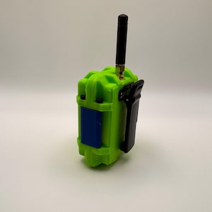 May include: A lime green electronic device with a black antenna and a black clip. The device has a blue rectangular panel and is likely a handheld radio or similar communication tool. The device is on a white background.
