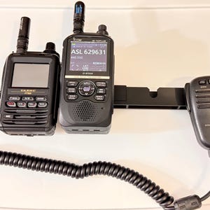 May include: Two-way radios and a microphone. The radios are black with antennas and screens displaying information. One radio has the text "YAESU FT60" and the other has "ICOM ID-51PLUS" with a screen displaying "ASL 629631". A black microphone is connected with a coiled cable.