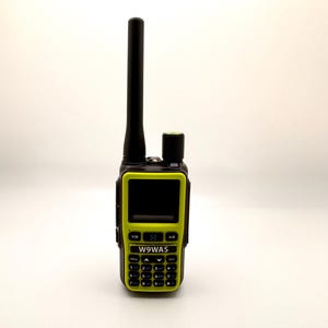 May include: A black and lime green handheld radio with a black antenna. The radio features a screen and a keypad with the text "W9WAS". The device is set against a white backdrop.