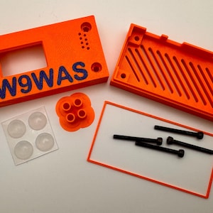 May include: An orange 3D-printed enclosure kit with the text "W9WAS" in blue. The kit includes two main orange pieces, a small orange component, a clear plastic sheet with four clear rubber feet, and several black screws.