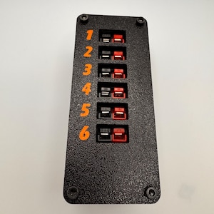 May include: A black rectangular panel with six pairs of black and red connectors. The numbers 1 through 6 are printed in orange. Four screws secure the panel. The panel has a textured surface.