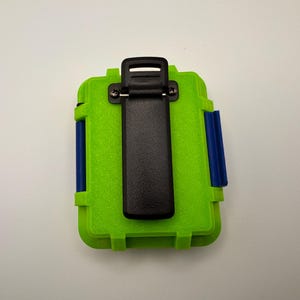 May include: A lime green rectangular case with a black clip on the top and blue accents on the sides. The case is designed to protect items and has a textured surface.