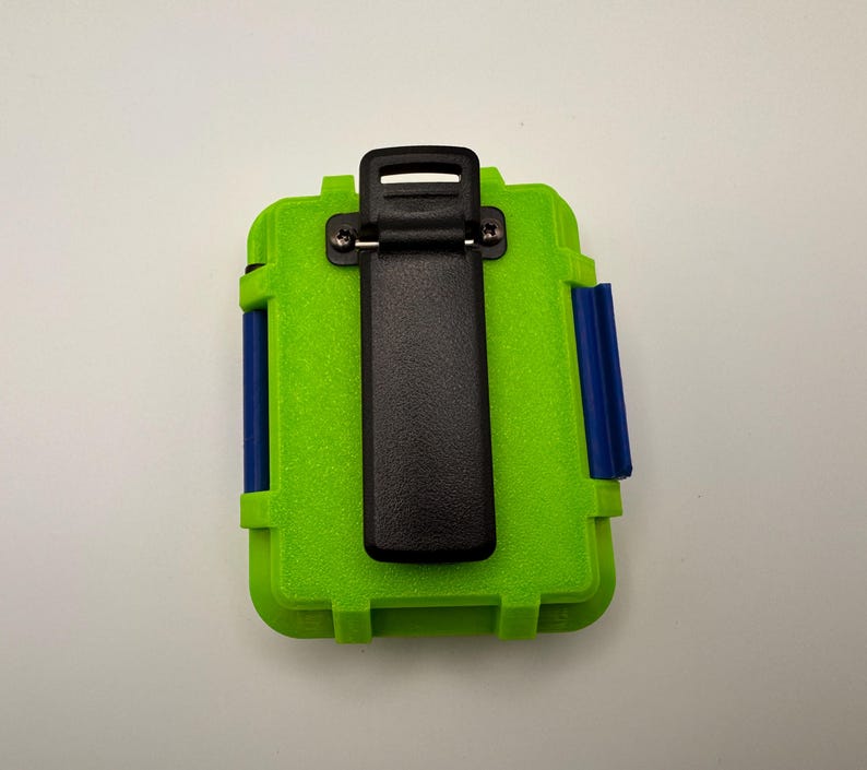 May include: A lime green rectangular case with a black clip on the top and blue accents on the sides. The case is designed to protect items and has a textured surface.