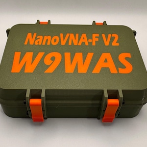 May include: An olive green case with orange text that reads "NanoVNA-F V2" and "W9WAS". The case has orange latches and hinges. The case is closed and appears to be made of a durable plastic material.