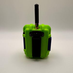 May include: A lime green electronic device with a black antenna and a black strap. The device has blue accents on the sides and a gold-colored connector for the antenna. The device is rectangular and appears to be a ruggedized enclosure.