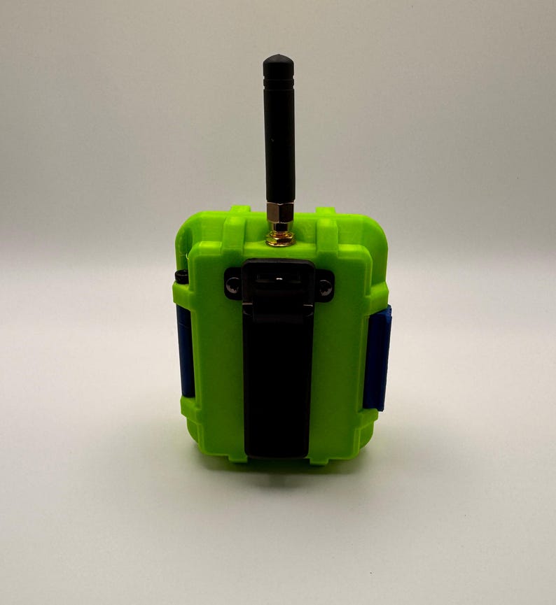 May include: A lime green electronic device with a black antenna and a black strap. The device has blue accents on the sides and a gold-colored connector for the antenna. The device is rectangular and appears to be a ruggedized enclosure.