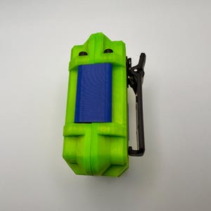 May include: A lime green rectangular device with a blue center panel and a black clip. The device has a textured surface and a geometric design. The clip is attached to the side of the device.