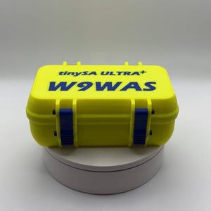 Könnte beinhalten: Eine leuchtend gelbe, rechteckige Box mit dem blauen Text "tinySA ULTRA+" und "W9WAS". Die Box hat blaue Verschlüsse und Scharniere. Die Box steht auf einer weißen Oberfläche.