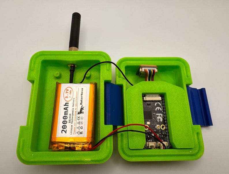 May include: Open green plastic case with a black antenna, a 2000mAh battery, and a circuit board. The case is open, revealing the internal components. The battery is labeled "MakerNova" and "3.7V". Wires connect the components.