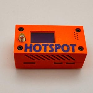 May include: An orange electronic device with the word "HOTSPOT" in blue lettering. The device has a small screen, a gold-colored antenna connector, and several small screws. The bottom of the device has a series of horizontal slots.
