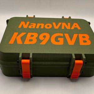 May include: An olive green case with orange text that reads "NanoVNA KB9GVB". The case has orange latches and hinges. The case is closed and appears to be made of a durable material.