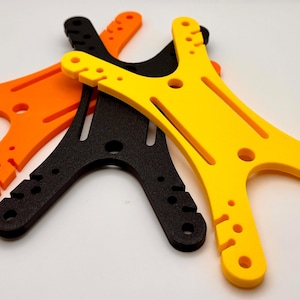 Peut inclure: Trois composants en plastique en forme de Y, de couleurs orange, noir et jaune. Chacun comporte plusieurs trous et fentes le long de ses bras et un trou central circulaire. Ils sont probablement destinés à un usage mécanique ou structurel.