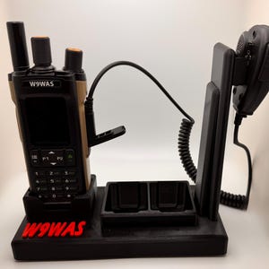 May include: Black radio communication device with a charging station. The radio is black and tan with a keypad and antenna. The charging station has two charging slots and a holder for a microphone. The text "W9WAS" is in red.