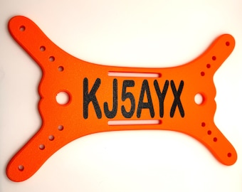May include: Bright orange license plate-shaped item with the text "KJ5AYX" in black. The item has a unique shape with extended ends and multiple holes. The background is plain white.