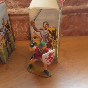 May include: A plastic toy knight figurine holding a shield with a red bird design. The figurine is standing on a small, brown base. The knight is wearing red and green armour and has a green helmet. The figurine is packaged in a cardboard box with a picture of a knight on it.