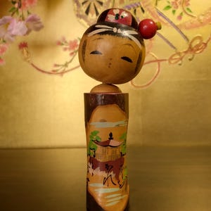 May include: A wooden Japanese Kokeshi doll with a painted face and a cylindrical body. The doll's head is round with closed eyes and a small red ball on the side. The body features a painted landscape scene with a temple.