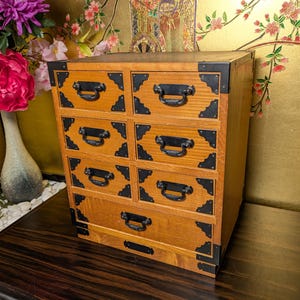 May include: A wooden chest of drawers featuring seven drawers arranged in four rows. The light brown wood cabinet is accented with black metal corner plates and black curved drawer pulls. The unit has a rectangular shape with a flat top surface.