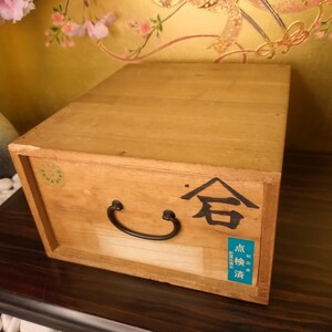 May include: A wooden box with a black handle and Japanese characters. The box is light brown with a rectangular shape. A blue and white sticker is on the front. The box is sitting on a dark brown surface.