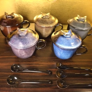 以下が含まれることがあります： 蓋とスプーンが付いた、6つのセラミックティーカップのセット。ティーカップは、茶色、グレー、青、紫など、さまざまな色があります。各蓋には、蝶の形をした装飾的なハンドルが付いています。スプーンは黒です。