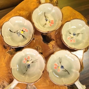 以下が含まれることがあります： 金色の縁取りが付いた、5つの小さな薄緑色の花型のセラミックボウル。各ボウルには、ピンク、黒、青で手描きされた花の模様があります。ボウルは木製の表面に配置されており、素朴な美学を示しています。