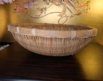 May include: A round, woven bamboo basket in light brown. The basket has a reinforced rim made of bamboo strips and white twine. The basket is shallow and suitable for various uses. The basket's diameter is approximately 30 cm.