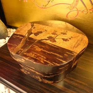 Puede incluir: Una caja de madera de forma ovalada con tapa. La tapa presenta un fondo dorado con un diseño de rama y flores. La caja tiene un exterior marrón oscuro texturizado. La caja está sobre una superficie de madera oscura.