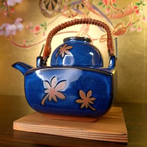 Könnte beinhalten: Blaue Keramik-Teekanne mit braunem geflochtenem Griff und Deckel. Die Teekanne hat braune Blattmuster und steht auf einem kleinen Holzsockel. Der Hintergrund ist goldfarben mit floralen Akzenten.