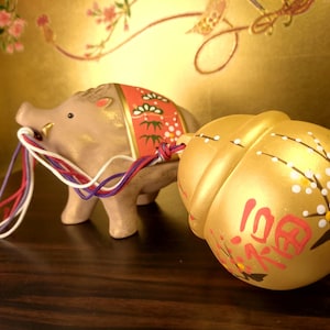 May include: A ceramic figurine of a tan wild boar with a red and green patterned saddle, pulling a gold-colored sphere. The sphere has Japanese characters and floral designs. The boar has colorful strings attached.