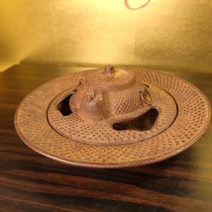Puede incluir: Un calentador de tetera redondo de hierro fundido de estilo antiguo con tapa. El calentador tiene una superficie texturizada con pequeños bultos espaciados uniformemente. La tapa tiene un asa pequeña.