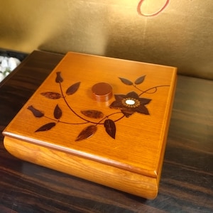 Op de afbeelding: Een houten doos met een bloemmotief op het deksel. De doos heeft een warme, middellange bruine kleur met een donkerbruine bloemeninleg. Het deksel heeft een centrale bloem en een kleine, ronde knop.