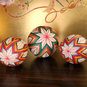 May include: Three decorative Temari balls, traditional Japanese folk art, are displayed. Each ball features a complex geometric design with red, yellow, green, and white thread. The balls are arranged on a dark wooden surface.