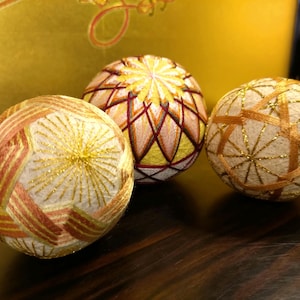 Puede incluir: Tres bolas Temari decorativas con intrincados patrones geométricos. Las bolas presentan hilos blancos, dorados y naranjas, creando una exhibición texturizada y colorida. El fondo es de un tono dorado, realzando los colores vibrantes de las bolas.