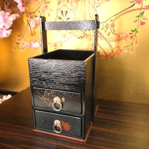Könnte beinhalten: Schwarze Holzschmuckschatulle mit zwei Schubladen und einem oberen Fach. Die Schubladen haben goldfarbene Ringgriffe und florale Designs. Die Box hat eine strukturierte Oberfläche und einen roten Rand.