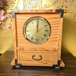 May include: A light-colored wooden table clock featuring a circular gold-toned face with black Roman numerals and thin hands. The rectangular frame has horizontal grooved detailing, a single storage drawer with a dark metal handle, and black corner brackets.