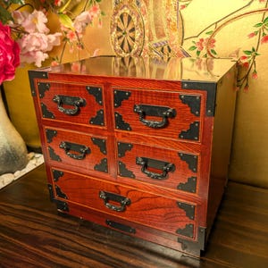 May include: A wooden chest with a reddish-brown wood grain finish featuring five drawers. Each drawer is equipped with black metal corner reinforcements and ornate black metal handles. The chest has a rectangular shape with a smooth top surface.