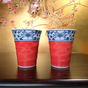 Puede incluir: Dos tazas de cerámica rojas y blancas con un diseño floral azul. Las tazas tienen un diseño texturizado y acanalado en la parte roja y un borde blanco con patrones florales azules. Las tazas están sobre una superficie de madera oscura.
