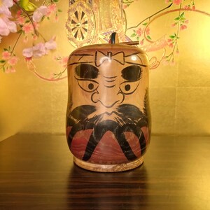 May include: A wooden Daruma doll with a painted face, featuring large black eyes, a mustache, and a beard. The doll has a brown body with red accents and a small black knob on top. The background is a golden color with floral accents.