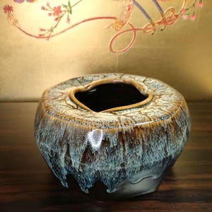 May include: A ceramic vase with an unusual, organic form. The vase has a dark interior and a textured exterior with a mix of brown, tan, and blue tones. The rim has a heart-shaped opening. The vase is on a dark wooden surface.