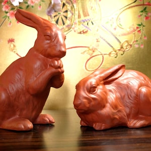 May include: Two terracotta-colored rabbit figurines. One rabbit is upright, with its paws together, and the other is in a seated position. The background is a golden color with floral and decorative elements.