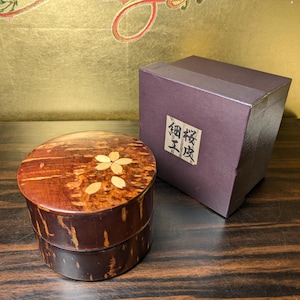 Puede incluir: Un recipiente redondo de madera marrón oscuro con textura de corteza natural y una incrustación floral clara en la tapa. Al lado hay una caja cuadrada de color púrpura oscuro con una etiqueta blanca que muestra los caracteres japoneses 桜皮細工.