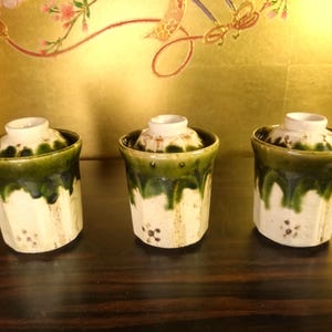 May include: Three ceramic containers with lids. The containers are white with a green glaze that drips down the sides. The lids are white. The containers are arranged on a dark wooden surface.