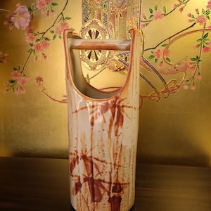 May include: A ceramic vase with a brown and white glaze and a bamboo design. The vase has a handle and is shaped like a cylinder. It is sitting on a wooden surface.