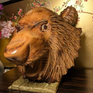 Peut inclure: Une sculpture en bois sculptée à la main représentant une tête d'ours avec une texture de fourrure détaillée, un museau lisse et une gueule ouverte. Le grain naturel du bois est visible sur toute la pièce, posée sur une base en tissu vert clair.