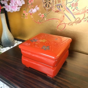 Puede incluir: Una caja lacada de color rojo vibrante, de forma cuadrada y acabado brillante. La caja está adornada con diseños florales en tonos azules, amarillos y grises. La caja está sobre una superficie de madera oscura.